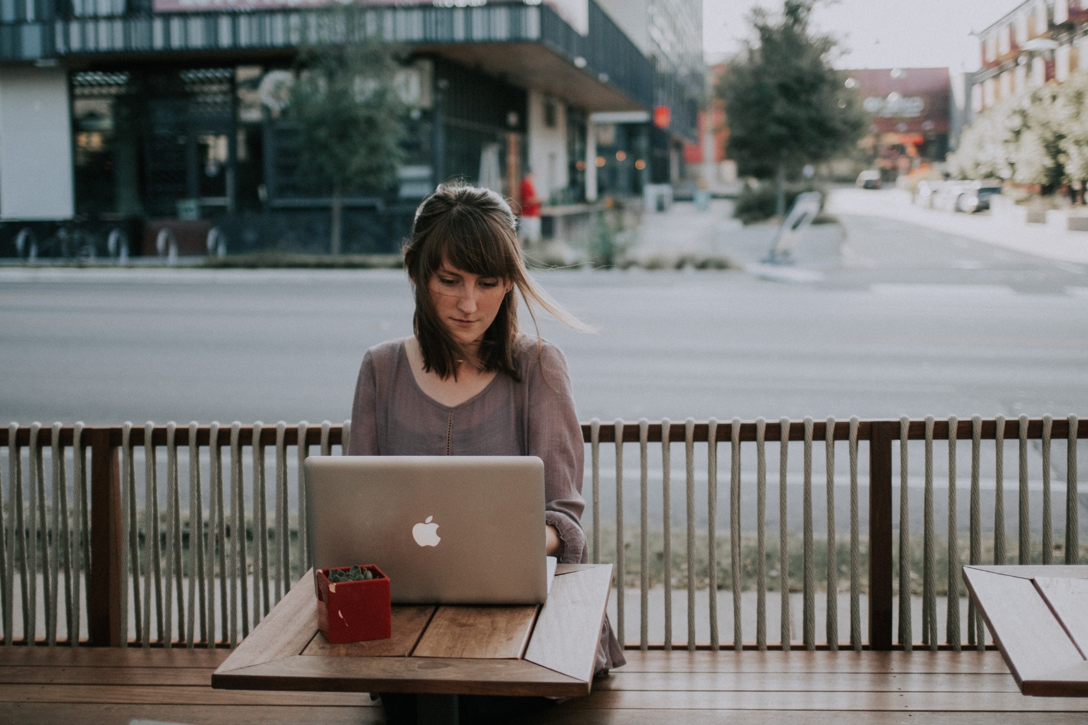 A woman sitting outdoors with her back to an empty city street. she has a mac laptop open in front of her.